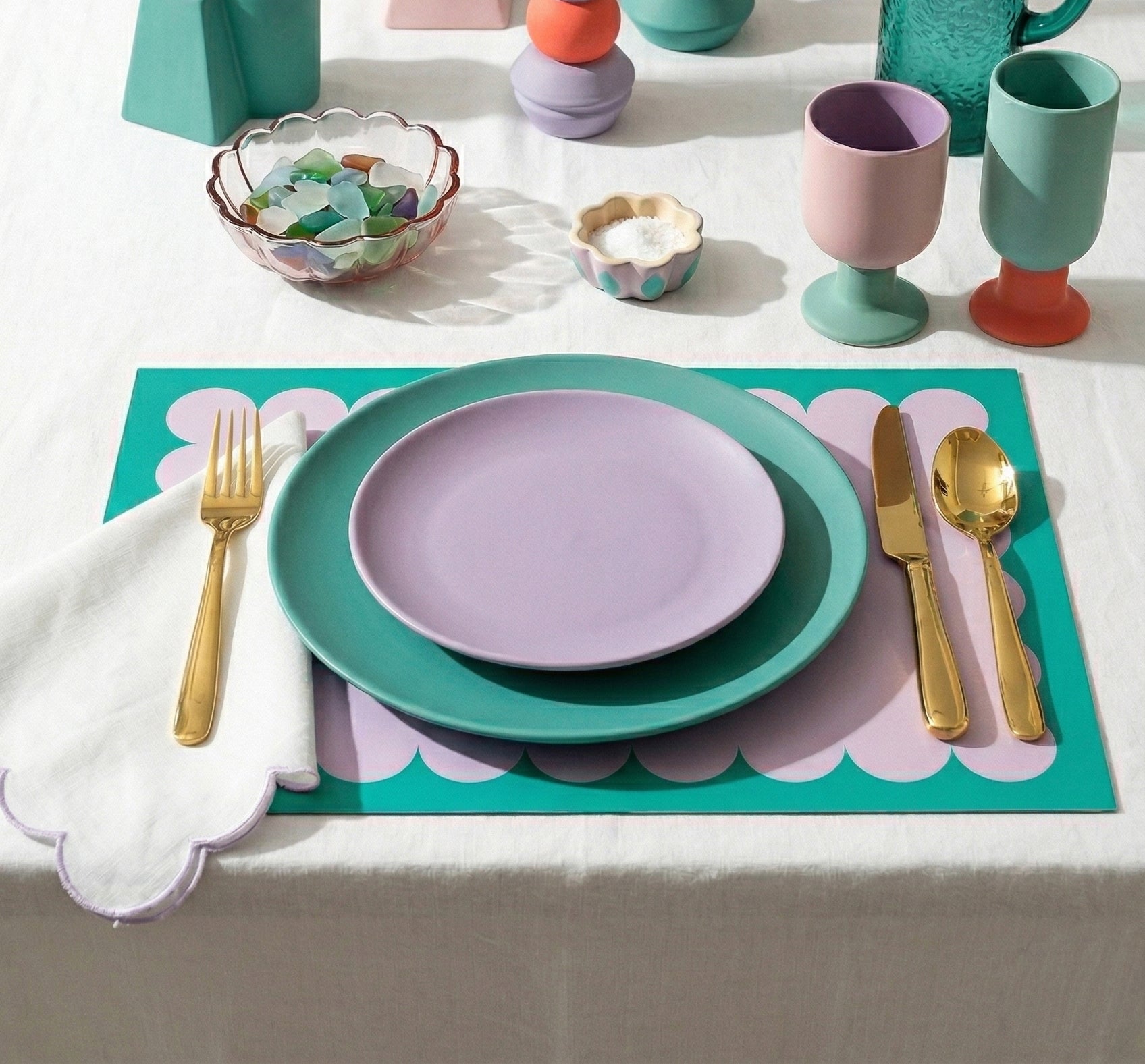 Colorful table setting with plates, cutlery, and scallop paper placemats in lavender and teal on a white tablecloth.