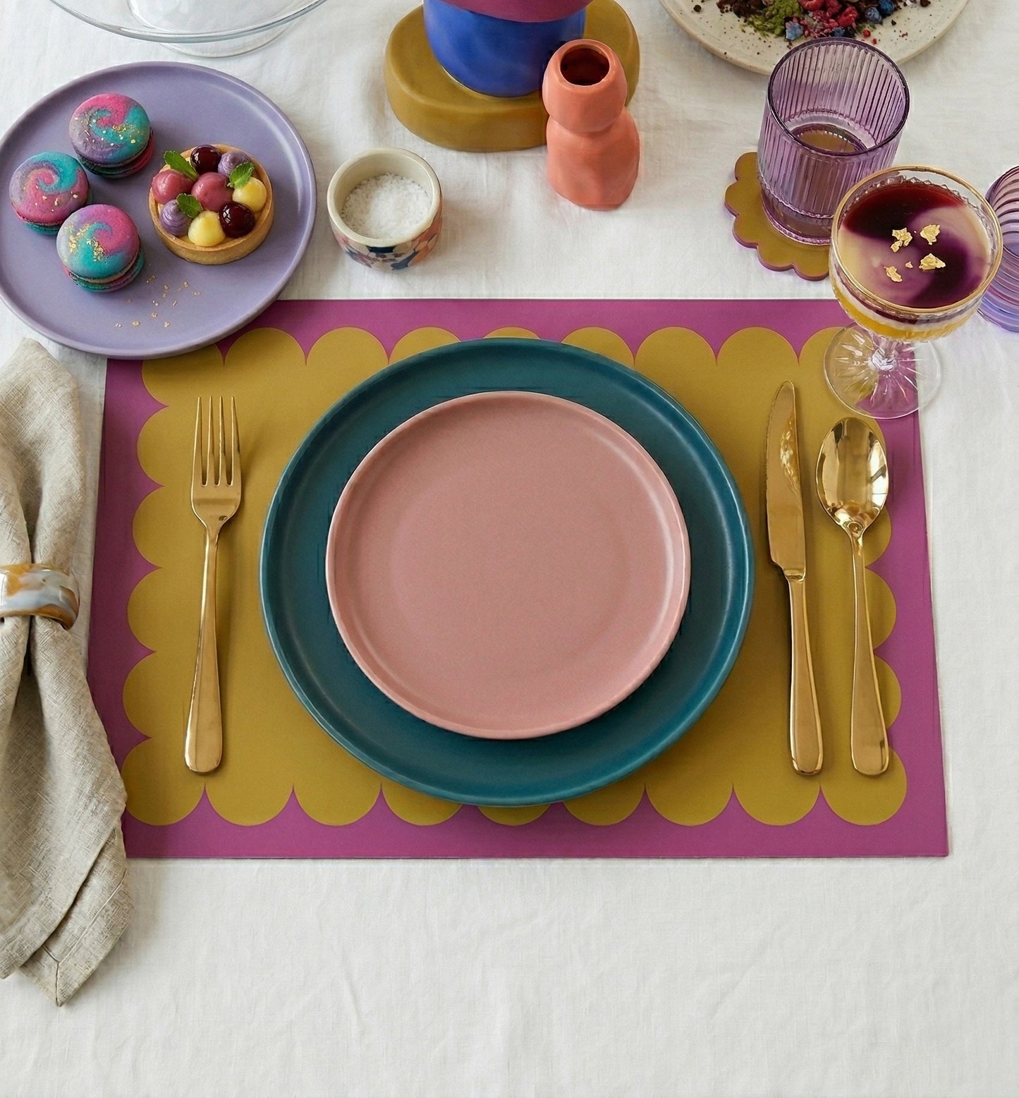 Colorful table setting with plates, cutlery, and glasses on a scalloped paper placemat in green and purple on a white tablecloth.