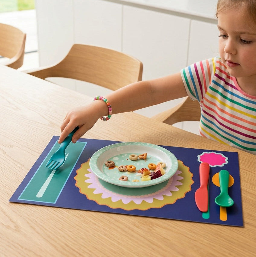 Child at a kitchen table with a plate of food and colorful utensils on a placemat.