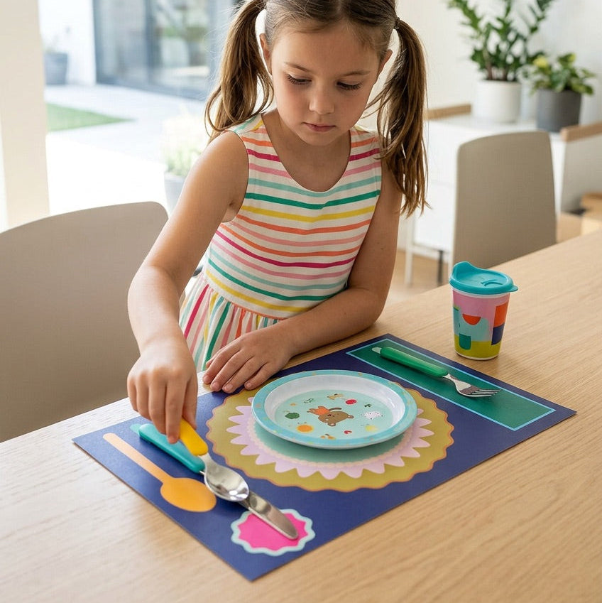 Young girl at a dining table with a paper placemat, plate, and utensils in a bright room.