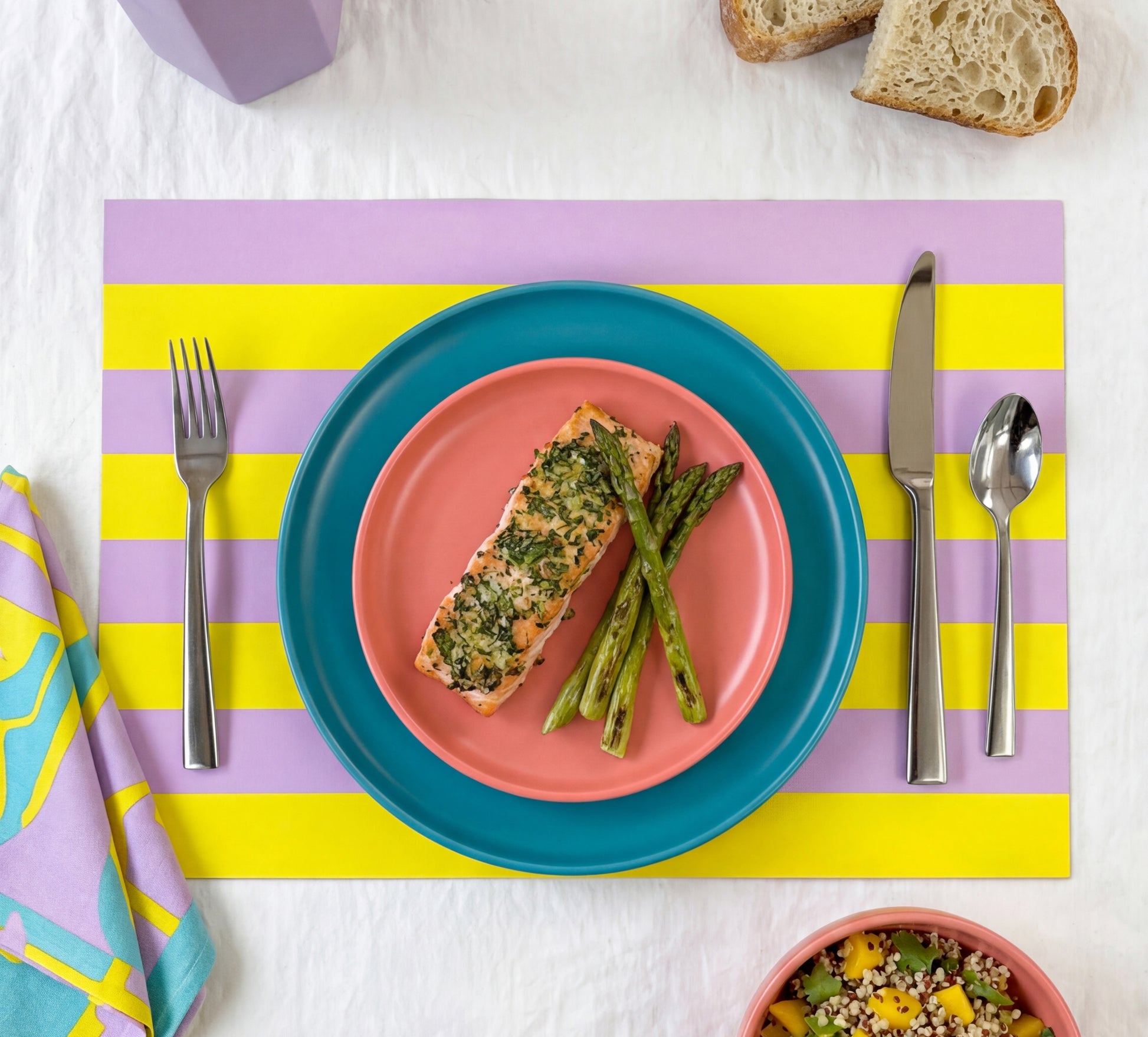 Colorful yellow and lavender striped paper placemat setting with salmon and asparagus on a pink plate, surrounded by cutlery and bread.