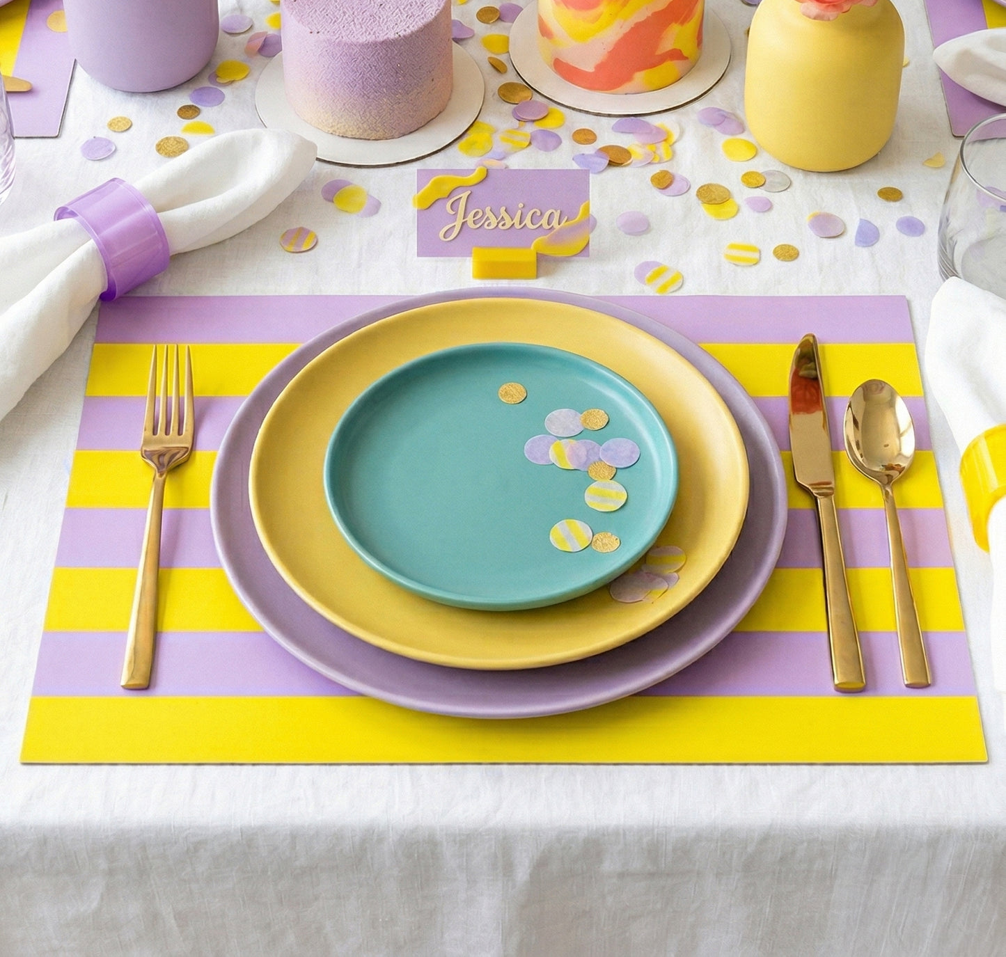 Colorful table setting with plates, cutlery, and yellow and lavender paper placemats on a white tablecloth.
