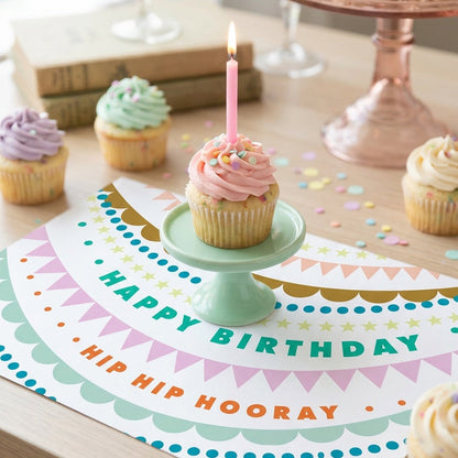 Birthday cupcake with a candle on a decorative placemat with 'Happy Birthday' text, surrounded by more cupcakes.