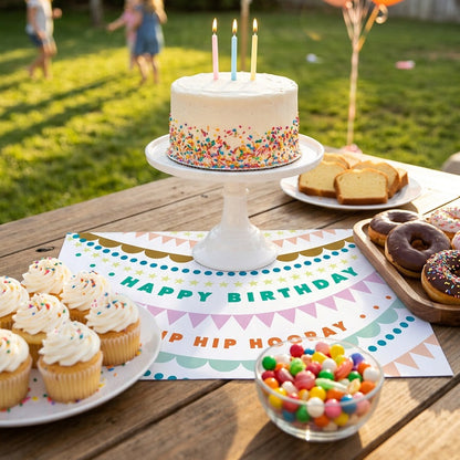 Birthday cake with lit candles on birthday paper placemat, surrounded by balloons and snacks.