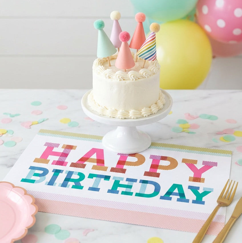 Birthday cake with colorful hats on a stand, surrounded by balloons and a 'Happy Birthday' paper placemat on a table.