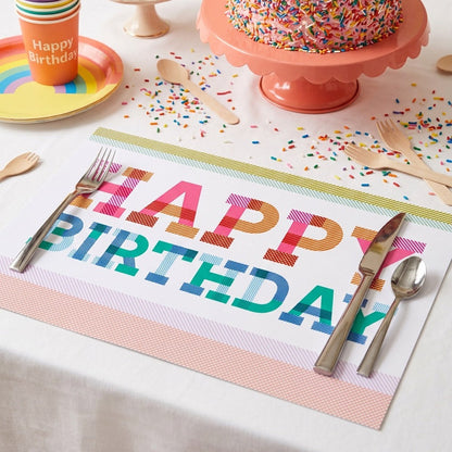 Birthday cake with candles on a pink stand, surrounded by party supplies including a 'Happy Birthday' paper placemat.