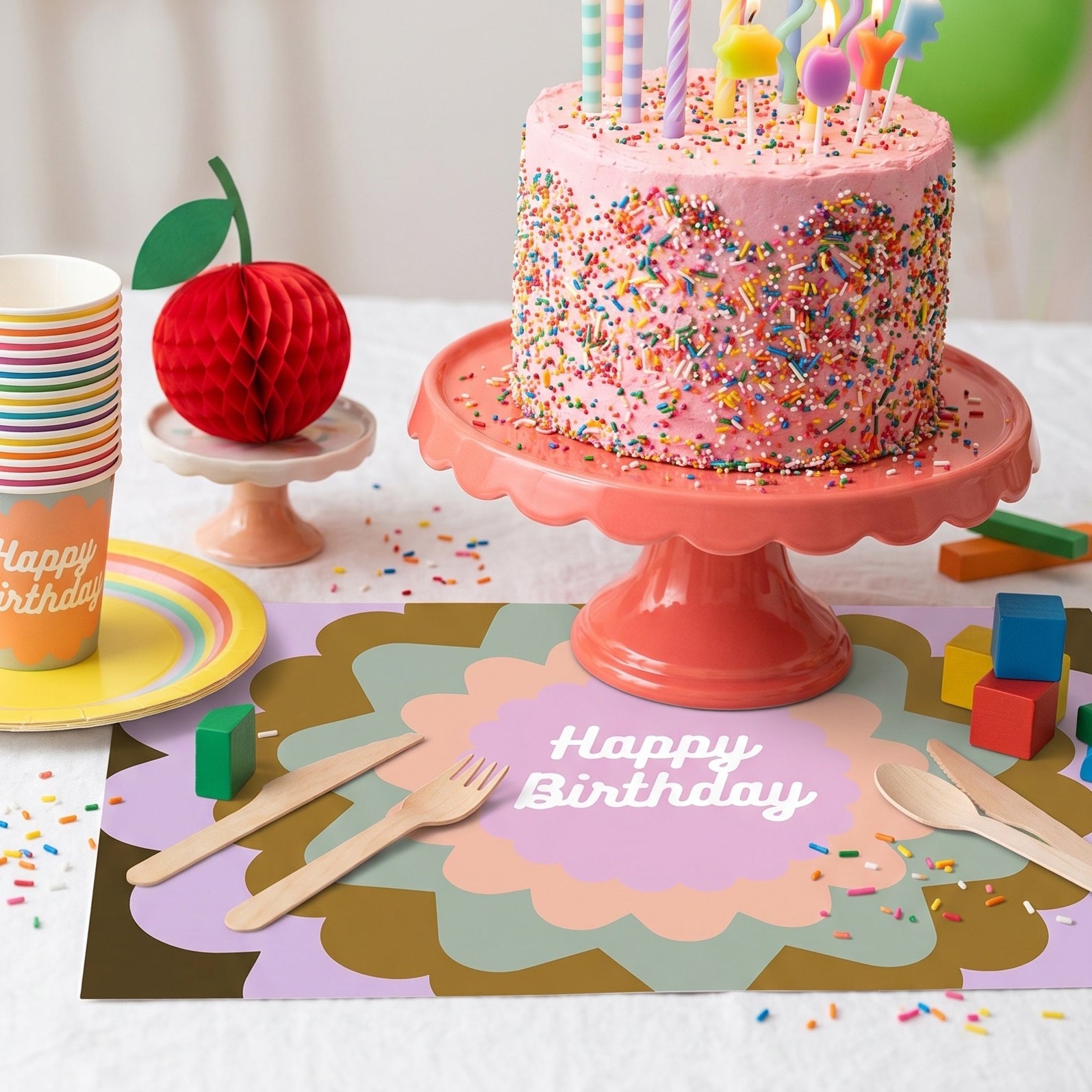 Birthday cake with lit candles on a pink cake stand, surrounded by happy birthday paper placemats and utensils.