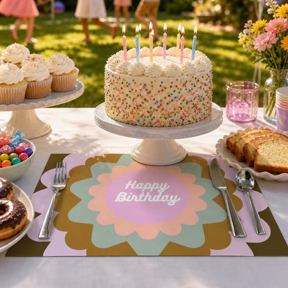 Birthday cake with lit candles on a birthday paper placemat outdoors with children in the background