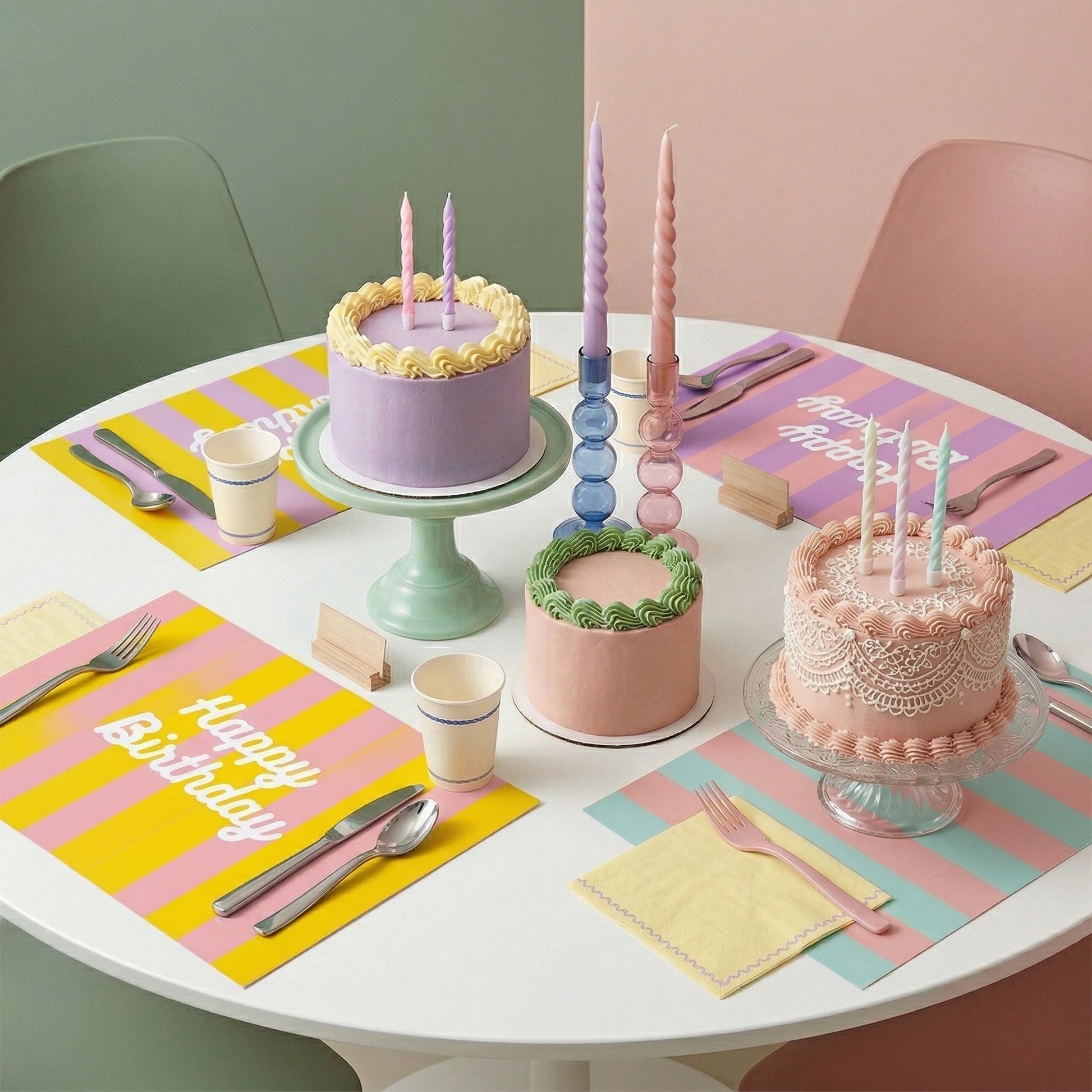 Round table with birthday cakes and colorful striped placemats on a pastel background