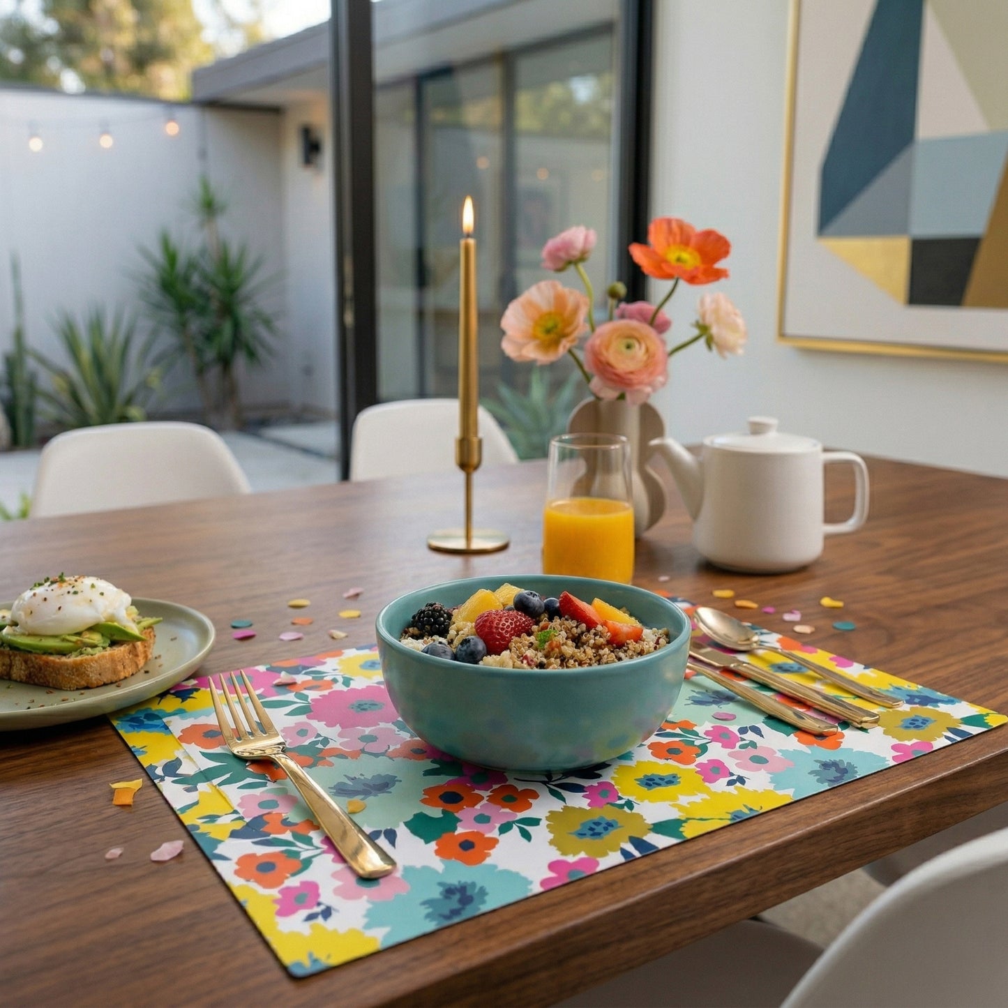 Dining table with a bowl of cereal, fruit, and toast on a floral paper placemat, with a candle and flowers in the background.