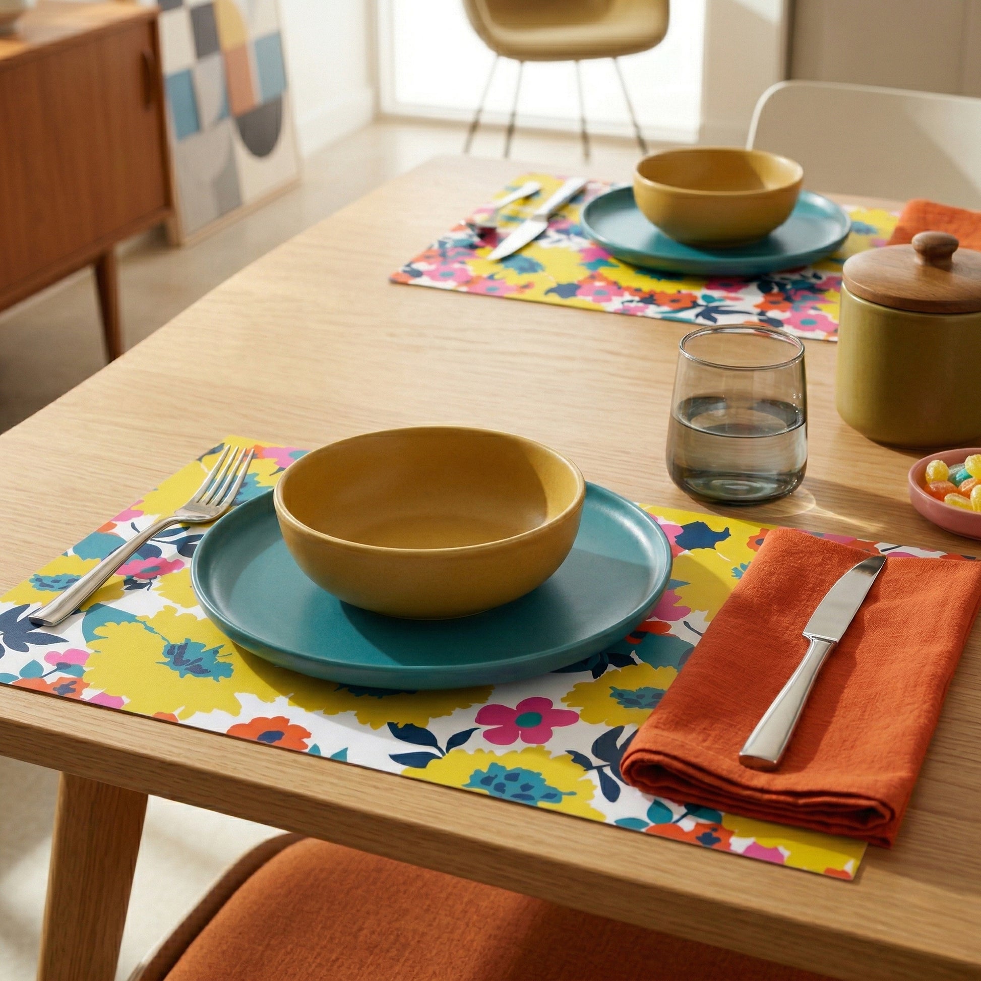 Dining table setting with colorful plates, bowls, and floral paper placemats.