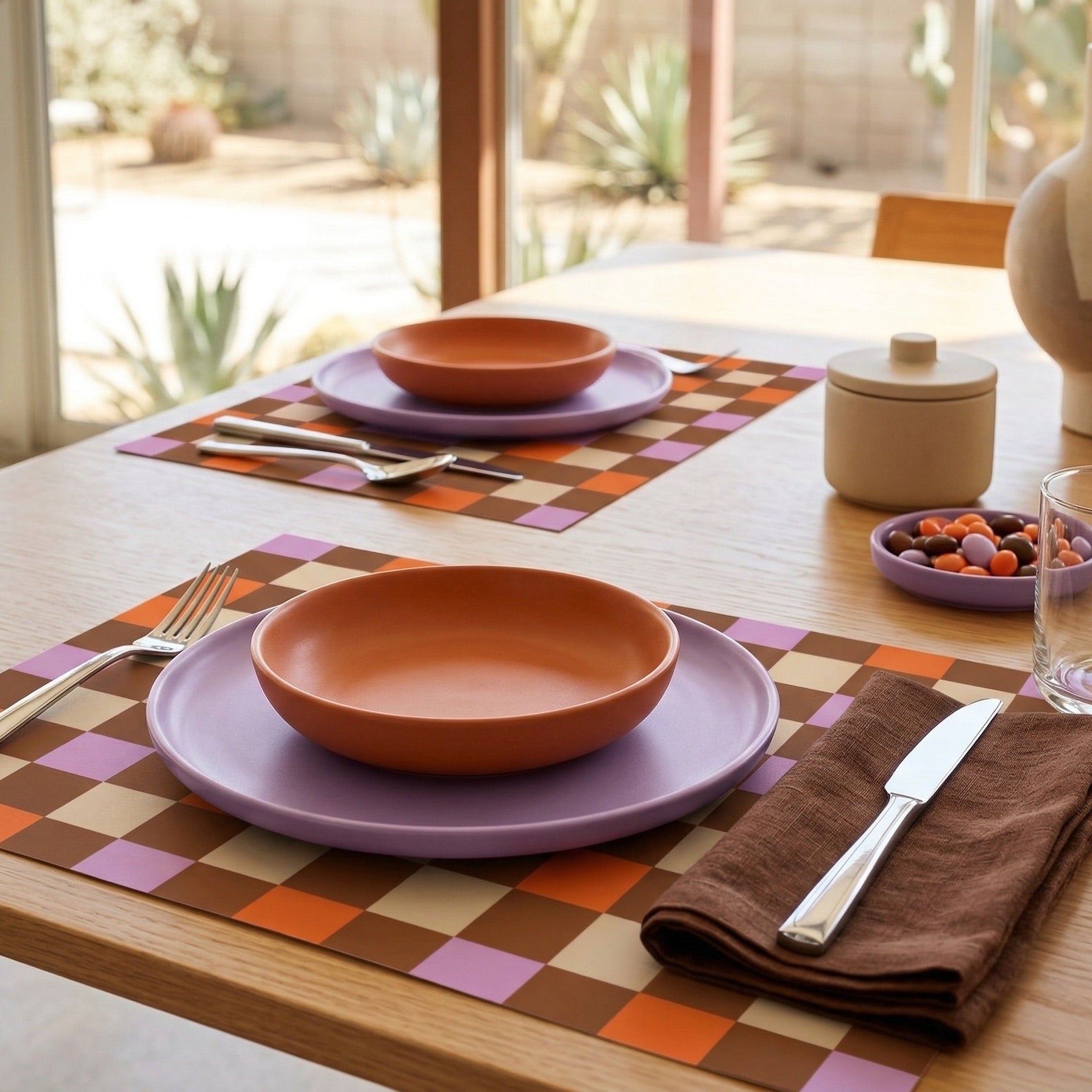 Dining table setting with orange bowls, purple plates, and a checkered paper placemat.