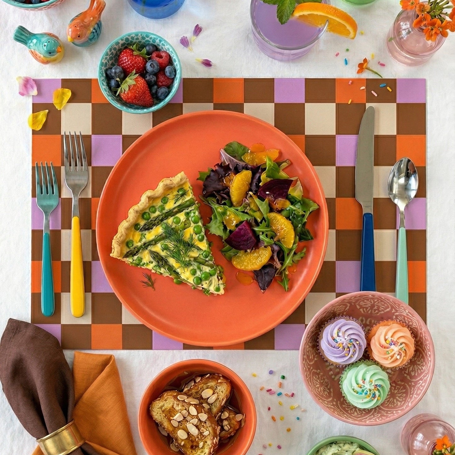 Colorful dining table setting with plates of food, flowers, and utensils on a checkered paper placemat.