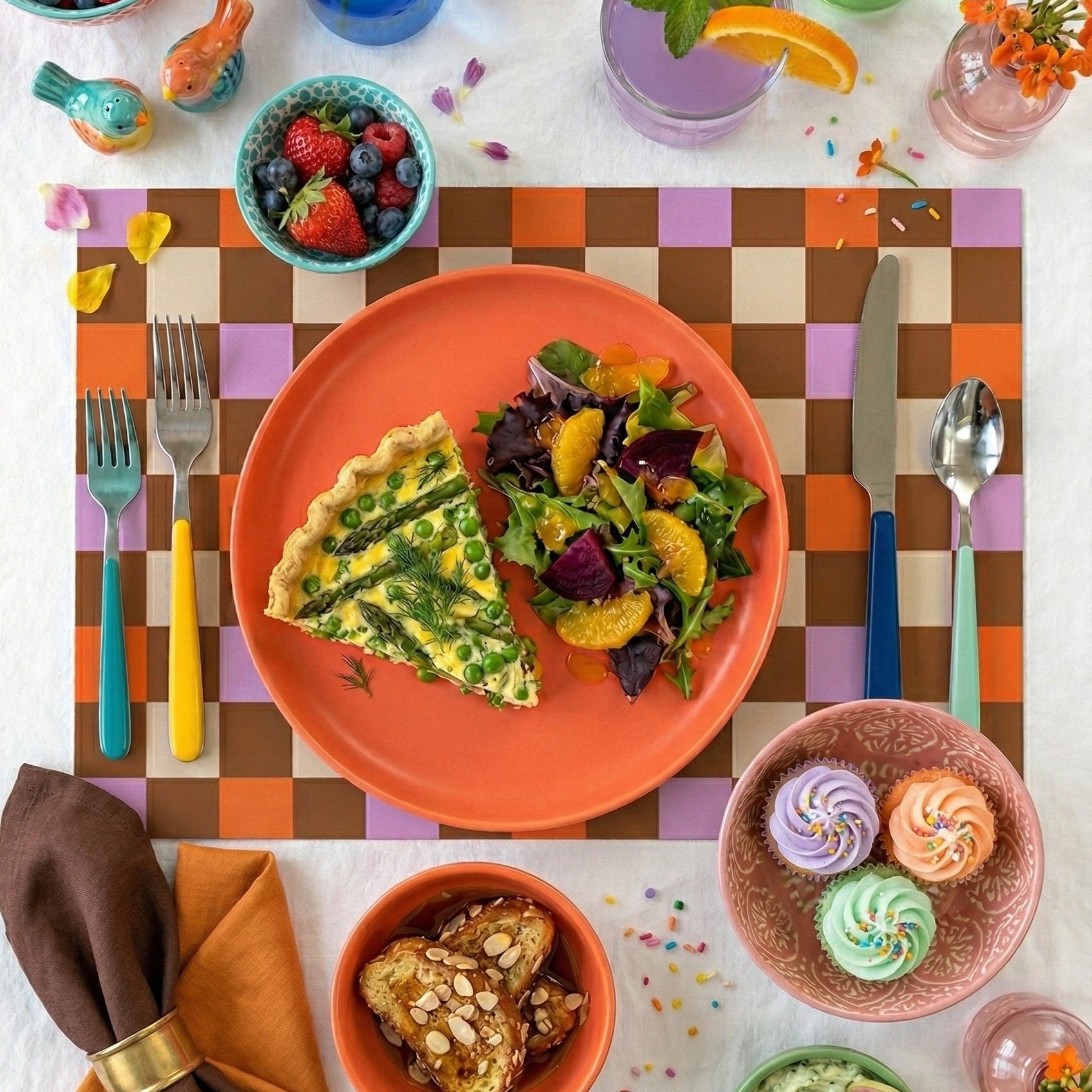Colorful dining table setting with plates of food, flowers, and utensils on a checkered paper placemat.