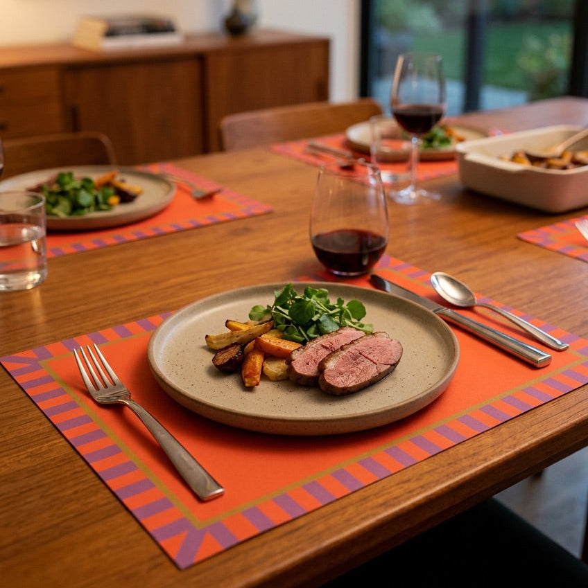 Dinner table setting with a plate of food, wine, and cutlery on a red paper placemat.