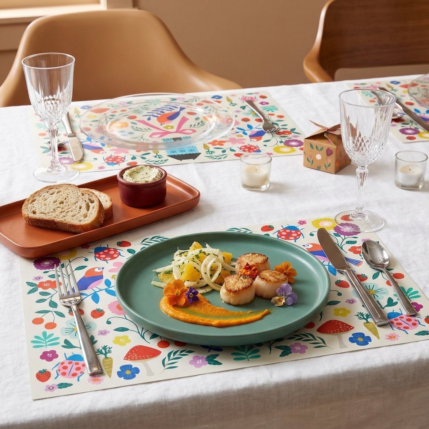 Colorful floral paper placemats styled on a table with scallops and bread.