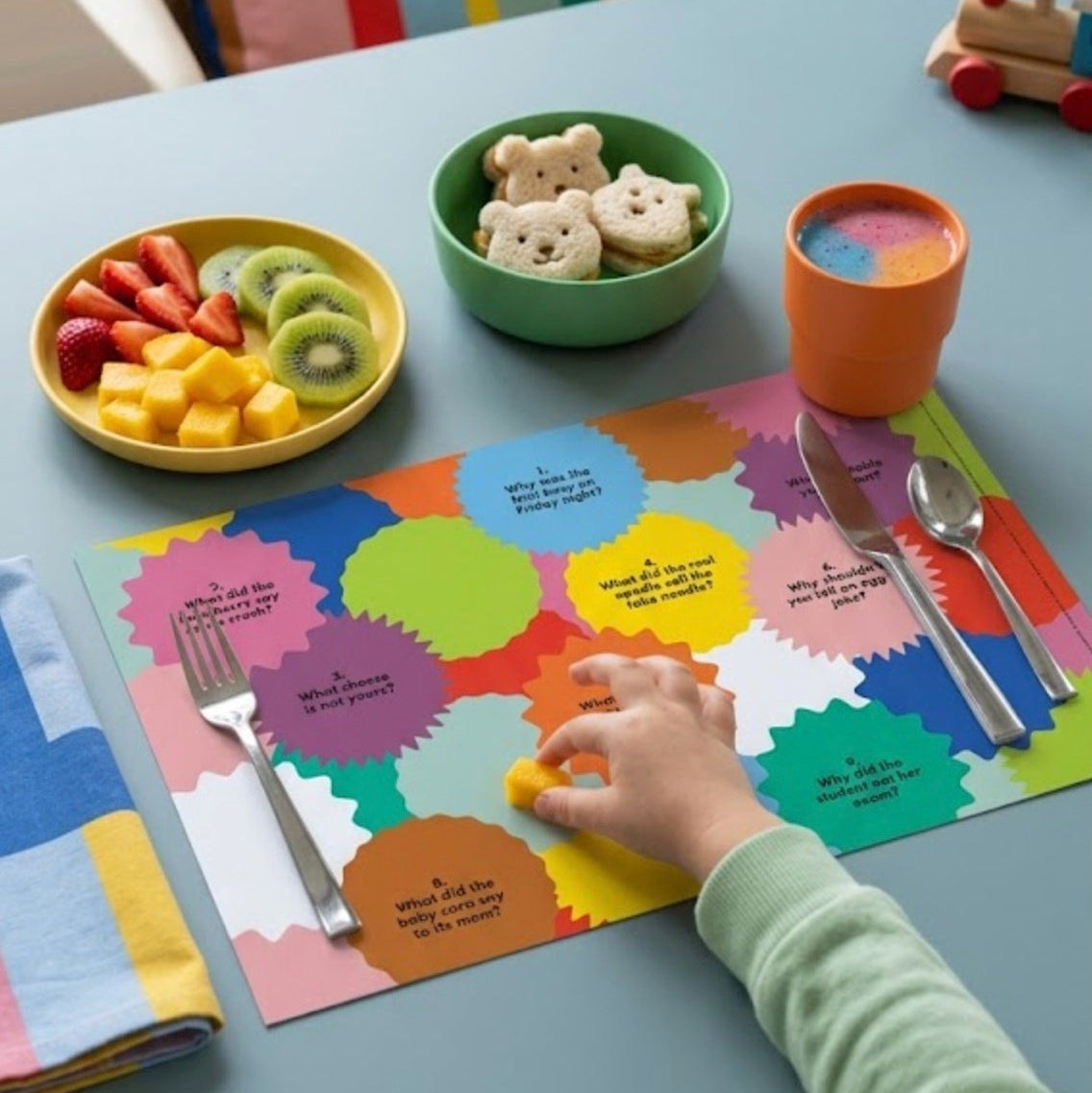 Child's hand reaching for a toy on a colorful paper placemat with food and utensils at a table.