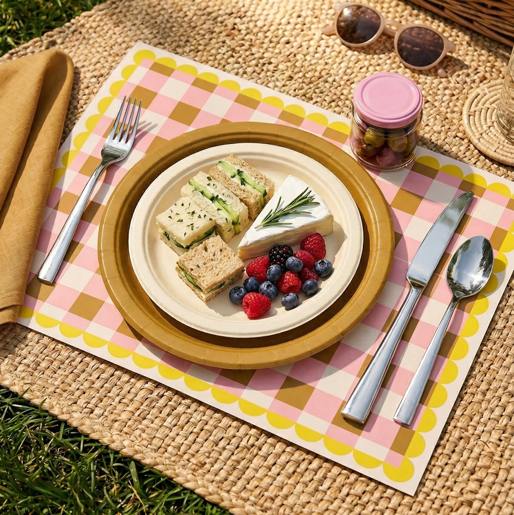 Picnic setup with a plate of sandwiches, fruit, and cutlery on a checkered paper placemat, with a picnic basket in the background.