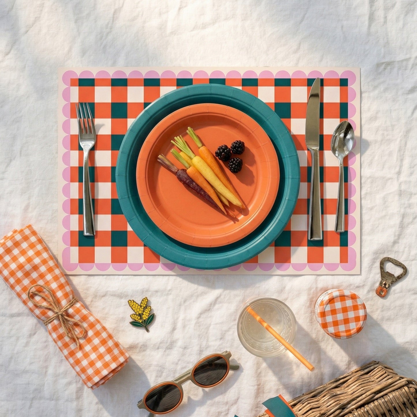 Outdoor dining setup with colorful plates, cutlery, and a checkered paper placemat on a white blanket.