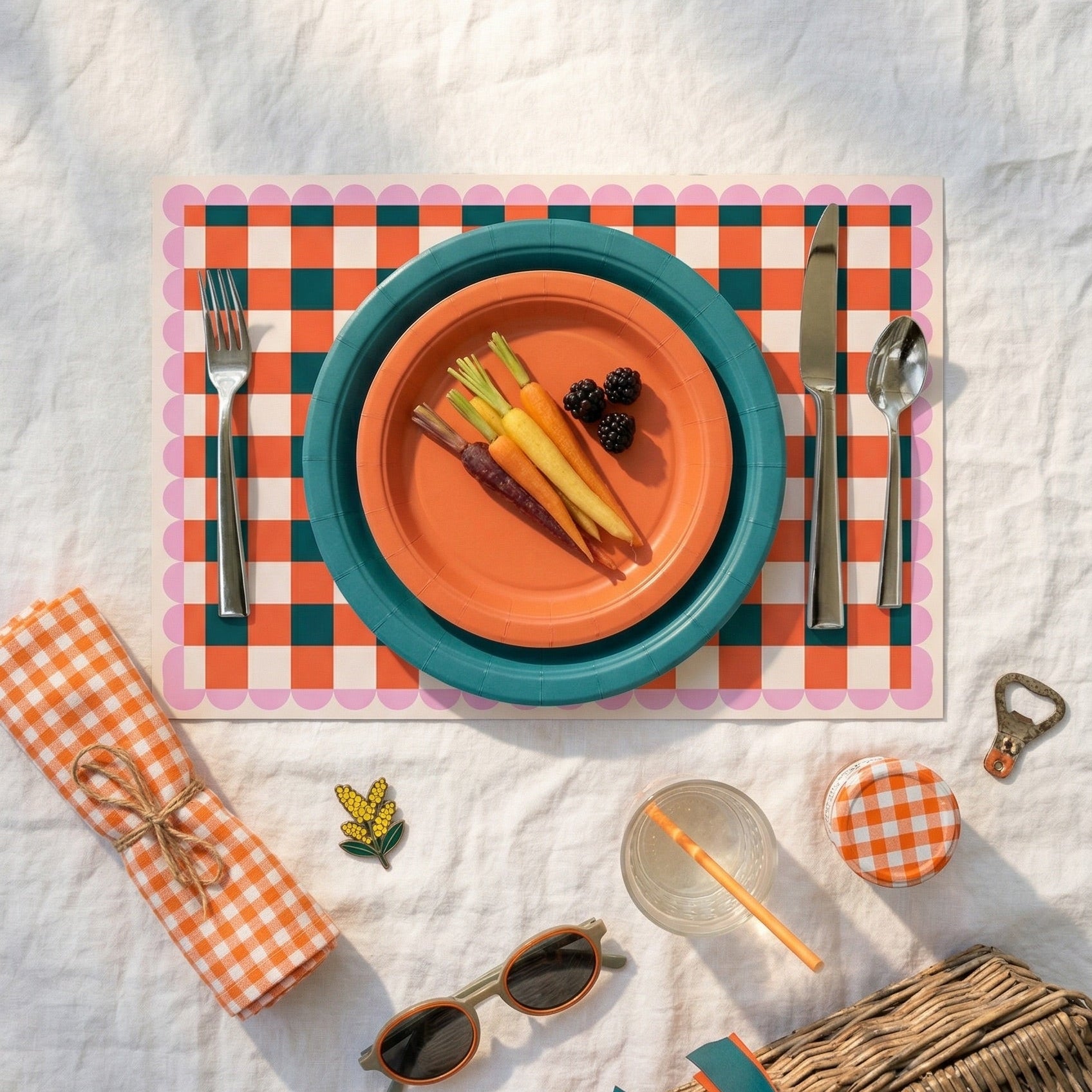Outdoor dining setup with colorful plates, cutlery, and a checkered paper placemat on a white blanket.