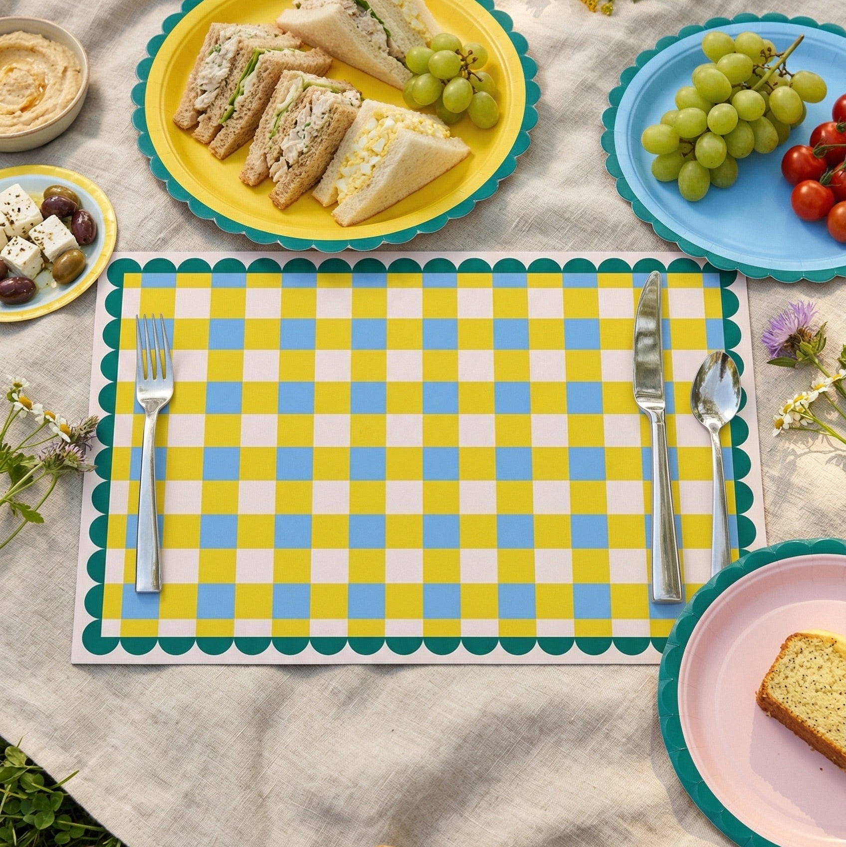 Picnic setup with plates of food, a checkered paper placemat, and a picnic basket on a blanket outdoors.