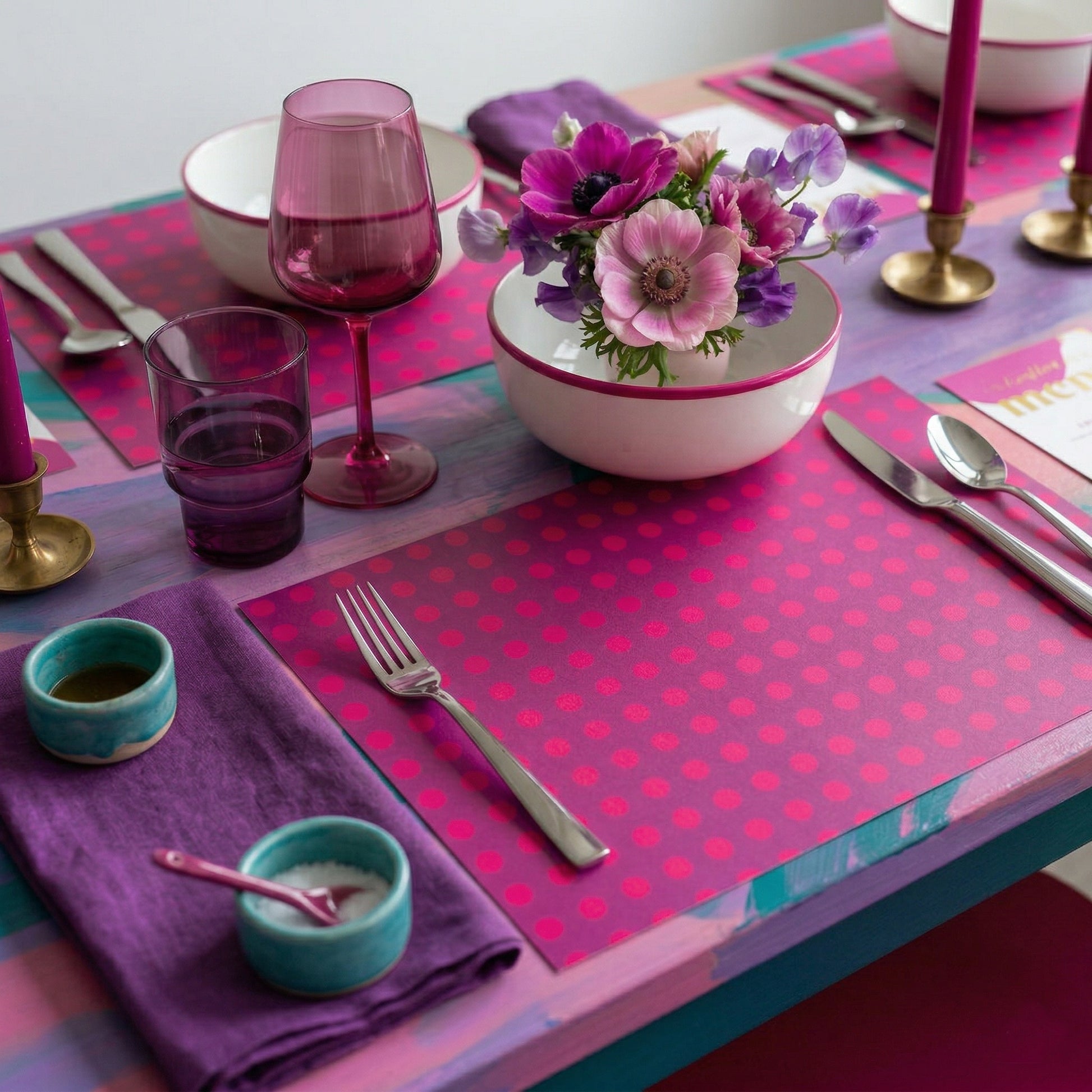 Colorful table setting with pink and purple paper placemats, including glasses, plates, and cutlery.