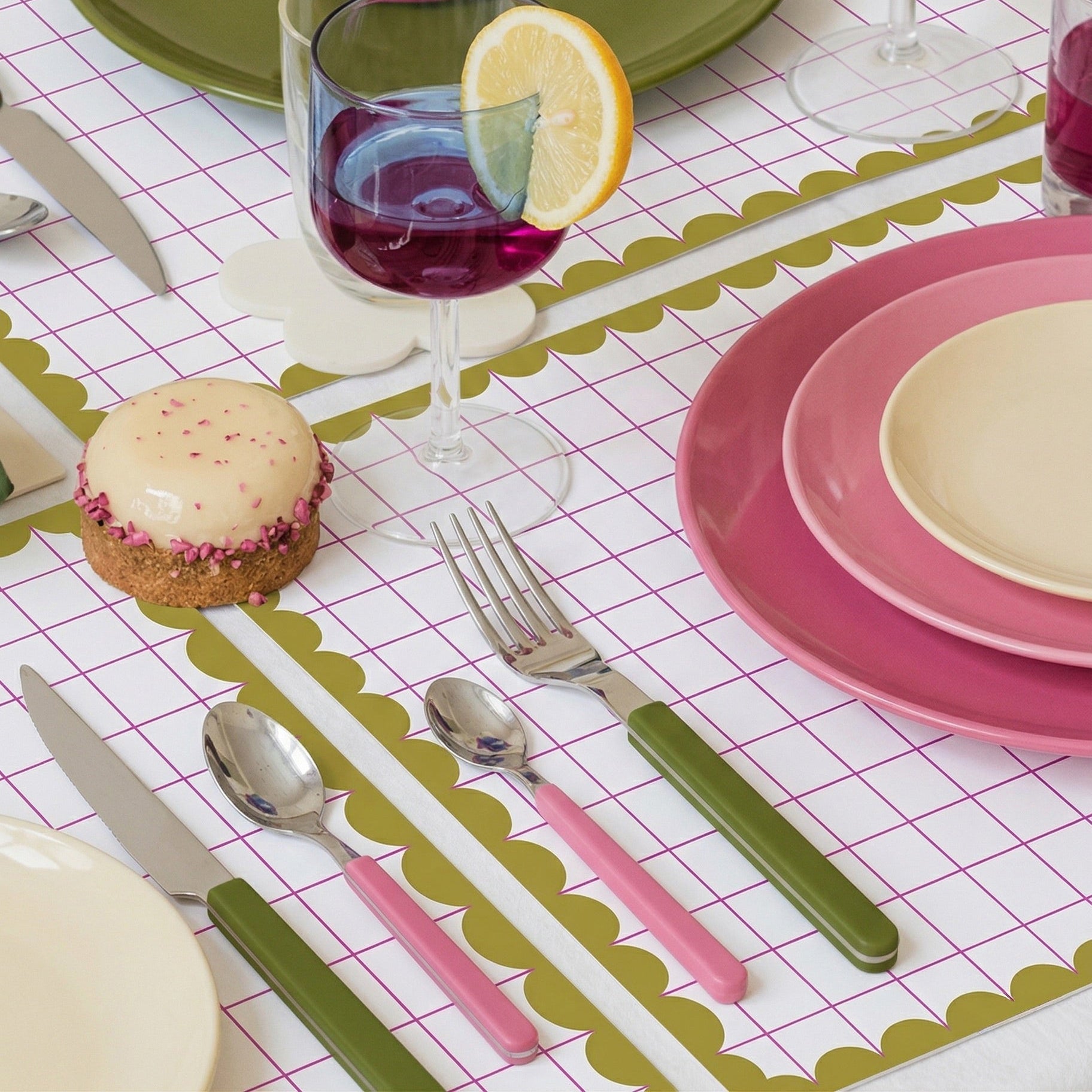 Dining table setting with pink and green plates, glasses, and utensils on a grid paper placemat.