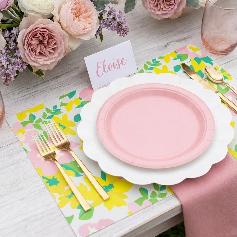 Table setting with pink plates, gold cutlery, and floral paper placemats.