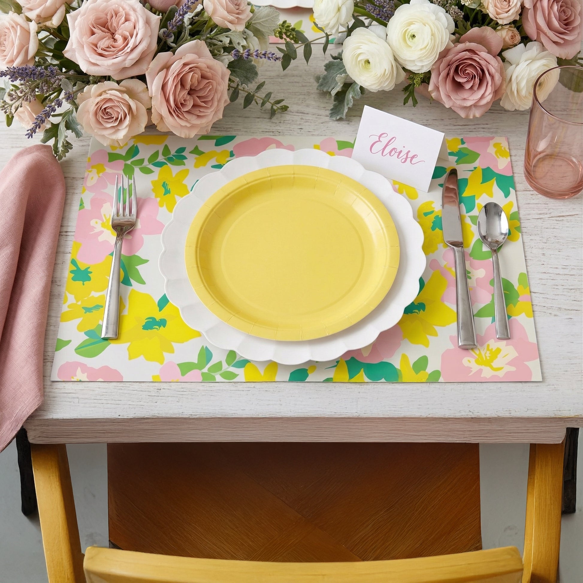 Table setting with a yellow plate, floral paper placemat, and pink roses.