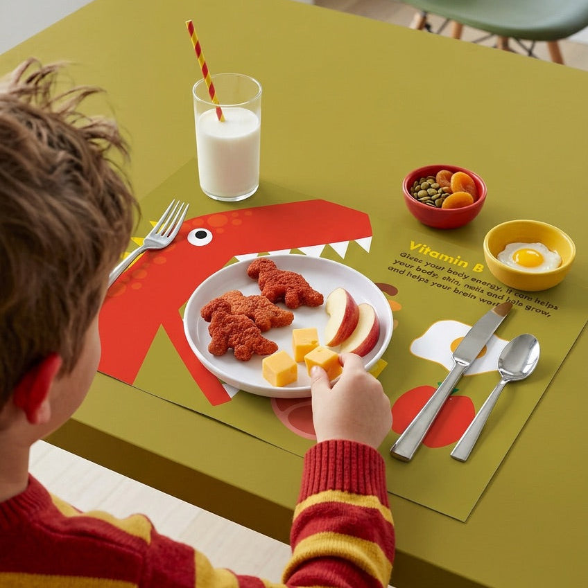 Child at a dining table with a dinosaur-themed paper placemat, eating a meal.