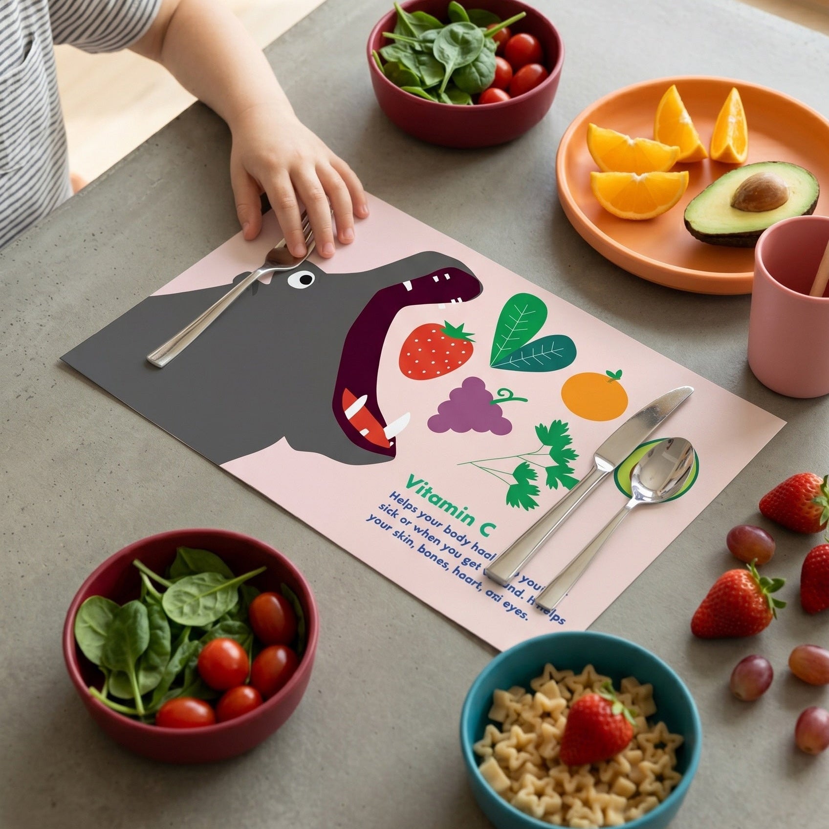 Children's mealtime setup with colorful plates, bowls of fruits and vegetables, and a placemat with cartoon illustrations on a table.